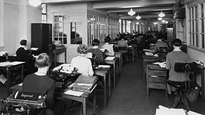 A room of women at typewriters. All face away from the camera.