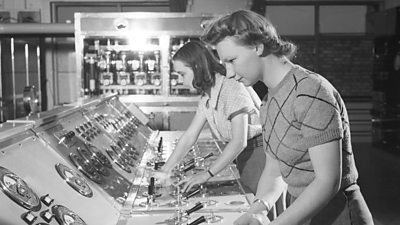 Two women concentrating on the controls of an enormous console with big dials and switches. 