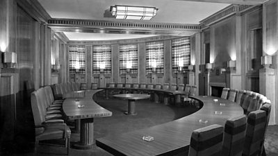 A large semi-circular room with wood panelling filled with three huge semi-circular tables set up for a meeting. There is nobody in the room. 