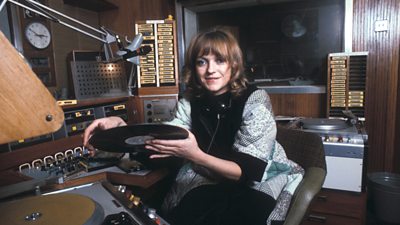 A young Annie Nightingale in a studio. She is holding a vinyl record in front of a deck. 