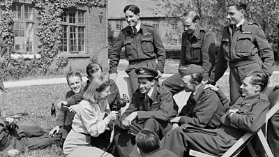 Noni Wright interviewing New Zealand airmen in uniform, relaxing in deckchairs the garden of the officer's mess. She is holding a tv microphone. 