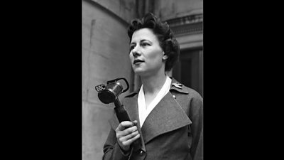 Mary Hill holding a showerhead-style tv microphone. Her jacket has enormous lapels.