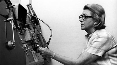 Peggie Broadhead, wearing heavy glasses, concentrates on operating a huge reel-to-reel tape machine. 