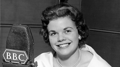 A young Judith Chalmers smiles broadly behind a tv microphone.