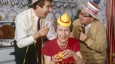 Zena Skinner seated peeling an orange wearing a party hat, while Leslie Crowther and Peter Glaze lark about behind in union jack hats.