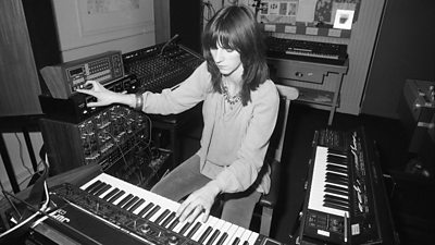 A woman at a keyboard while turning a multitrack recording switch with the other hand.