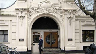 A white fronted building a round arched door with ornate details. A man walks past on a mobile phone. 