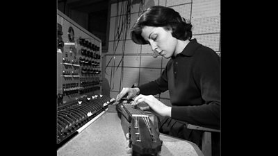 A woman in front of an audio modulator plays a stringed instrument on the table. 