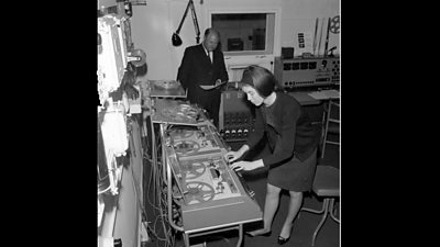 A woman stands in front of a bank of reel-to-reel tape machines.