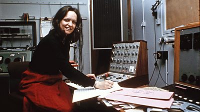 A woman posing at a 1970s synthesizer. She is writing on music manuscript paper.