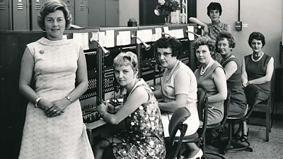 Women telephone operators seated at their equipment, which have large fixed microphones. All wear dresses, many with pearls. 