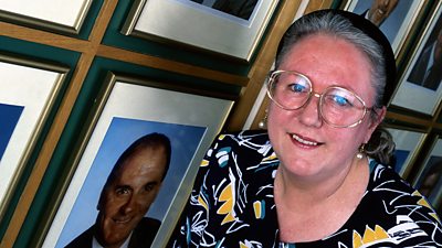 Frances Line stands in front of a vast wall of corporate photographs of men.