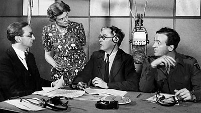 A woman standing discusses a script with three seated men. A lozenge shaped tv microphone hangs from the ceiling. 