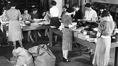 An industrious scene - 11 women and a 1 teenage boy sort the post at two large tables. Sacks of mail await on the floor. 