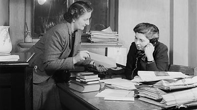 A woman on the telephone is shown a letter by another. Her desk is full of newspapers and books. 