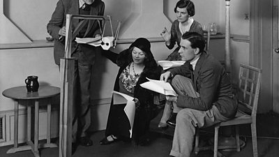 Two men and two women gathered around a microphone with scripts in hand. 