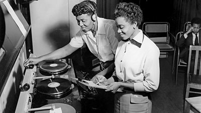 Two women at the gramophone, one supervising, the other operating the machine.