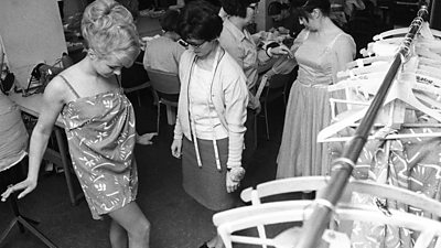 Two women being measured by wardrobe staff with tape measures. The women being dressed has a very 1960s beehive hairstyle. 