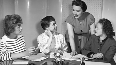 Three women gathered around the microphone. The female producer, standing, appears to have told a joke. 