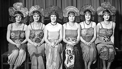 Six seated women smile at the camera. They are wearing dresses with extravagant halo-like headdresses.