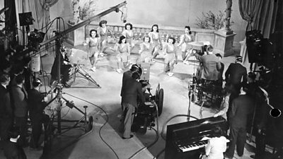 A television set in the 1940s - eight women dancing, around lit stage are three cameras, light and a boom microphone, plus a pianist.