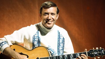 Wearing a white roll-top jumper and holding his guitar, Val Doonican smiles toothily at the camera.