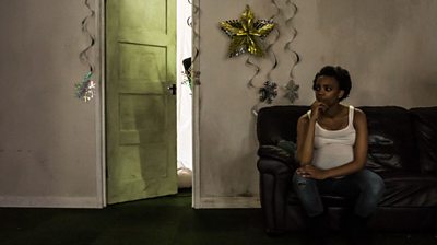 A woman sits alone in a shabby flat with Christmas decorations