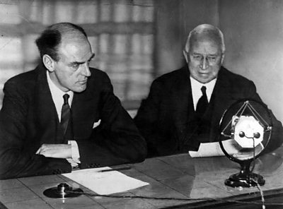 Two suited men at a desk with a 1930s microphone. Reith is on the left.