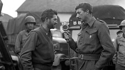 A soldier is interviewed by a 鶹ҳ journalist also in uniform, with a large 鶹ҳ microphone. Background is soldiers in front of large lorries.