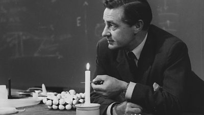 A man in a suit with a blackboard behind and a lit candle.