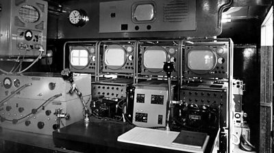 A bank of four monitors and various pieces of equipment with dials and switches.