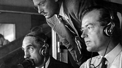 Two seated men in headphones, one with a commentator's microphone. Another man leans in. 