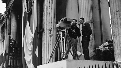 Two cameraman look through an early TV camera on a tripod.