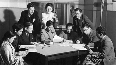 10 broadcasters of different nationalities read scripts around a table. A tv microphone hangs from the ceiling. 