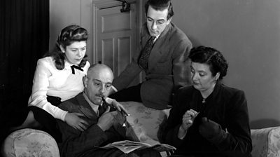 Two young adults look over the shoulder of their balding father reading a newspaper. He smokes a pipe, while his wife, knitting, looks on.