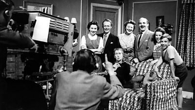 A BBC camera records a family gathered around a sofa in a studio set.
