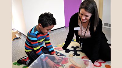 A woman therapist smiles as a little boy plays with toy cars. 