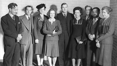 Men and women line up for a group photograph. All are dressed smartly in outfits of the 1940s.