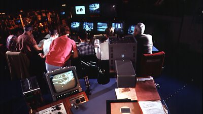 A TV production gallery - a mostly male crew watch a large bank of black and white TV screens. 