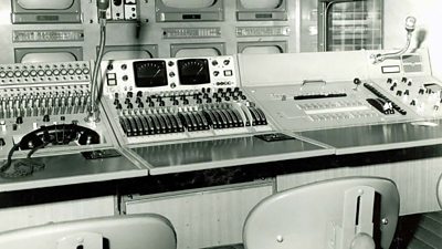 Interior of a TV outside broadcast van with a row of TV screens, various sliders and buttons on a panel and a desk with three seats. 