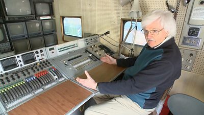 Brian Summers sits at the control desk with a large bank of TV screens and a communications panel with a microphone. 