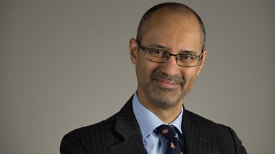 A man in a suit with glasses and a short stubble beard.