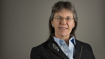 A blue suited woman with long hair, glasses and earrings. 