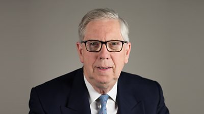 An older man with grey hair, glasses and a blue suit.
