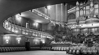 The interior of the Queen's Hall - a majestic sweeping balcony, with a large pipe organ. The stage has palm trees around the outside. 