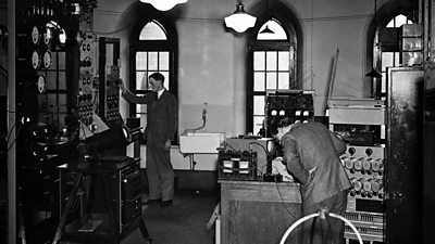 Two engineers in suits fiddle with various bits of analogue apparatus in a gothic room