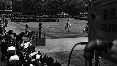 A large parade with soldiers in full number 1s takes place at Horseguards Parade - a soldier on a podium takes the salute. A ˿ TV camera looks on