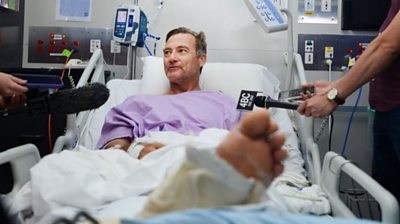 Neil Parker in a hospital bed surrounded by microphones