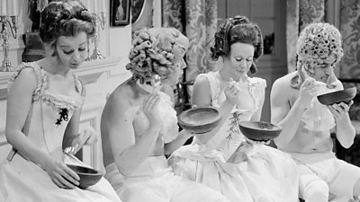 Terry Jones and Graham Chapman sit shirtless in regency wigs eating from wooden bowls. Two women look on disapprovingly