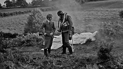 John Cleese as a parachutist is confronted in a field by Michael Palin as an Edwardian country gentleman with shotgun